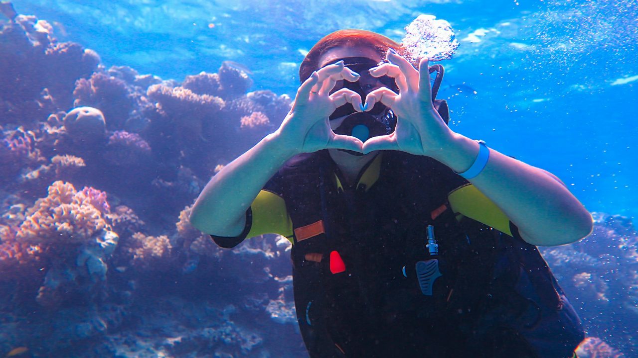 A girl in scuba diving near a sea reef, shows a heart with her hands